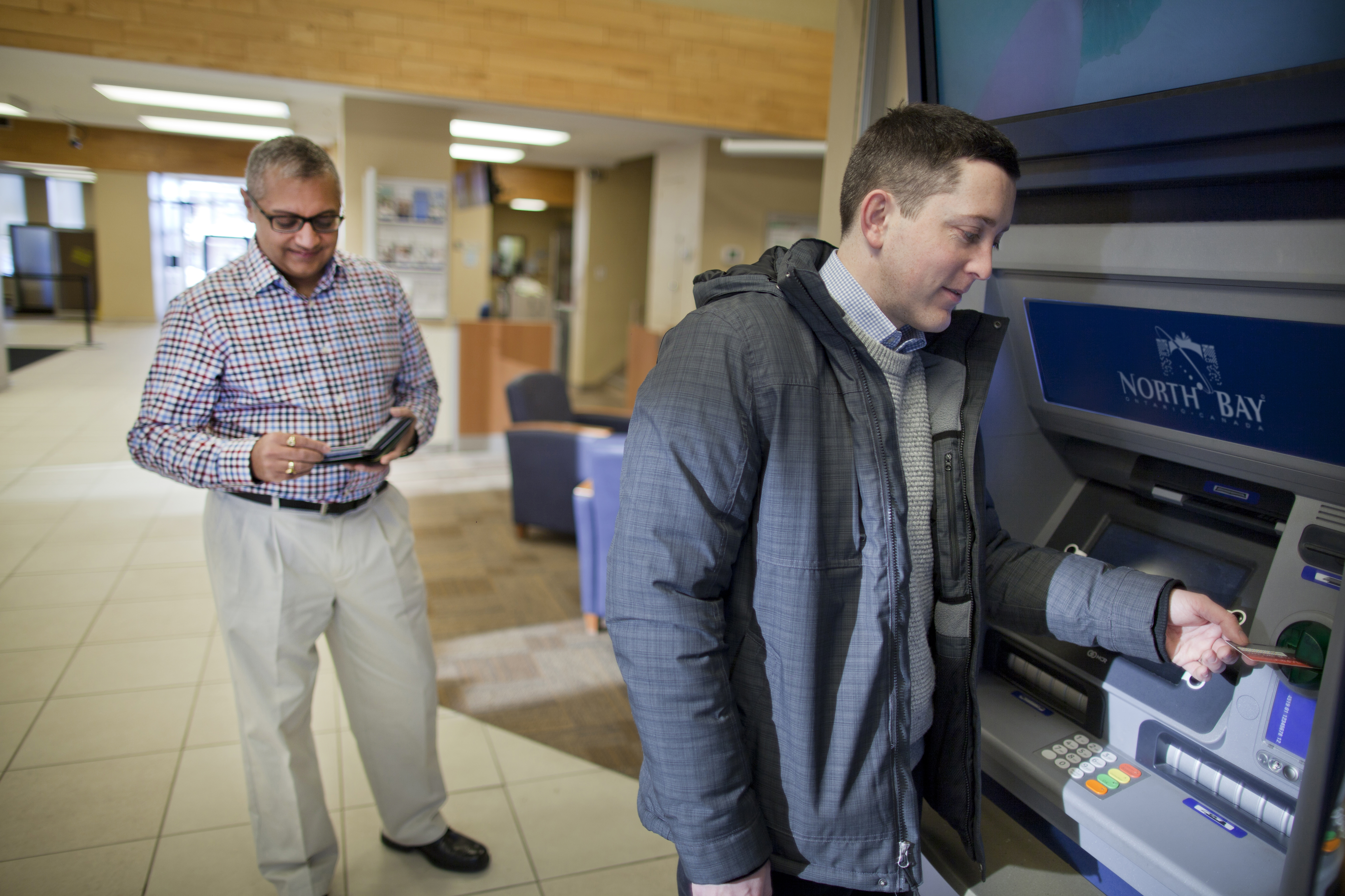 A man uses an ATM and another man waits in line.